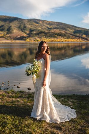 Stunning bride in lace gown poses by mountain lake at sunset holding white bouquet.