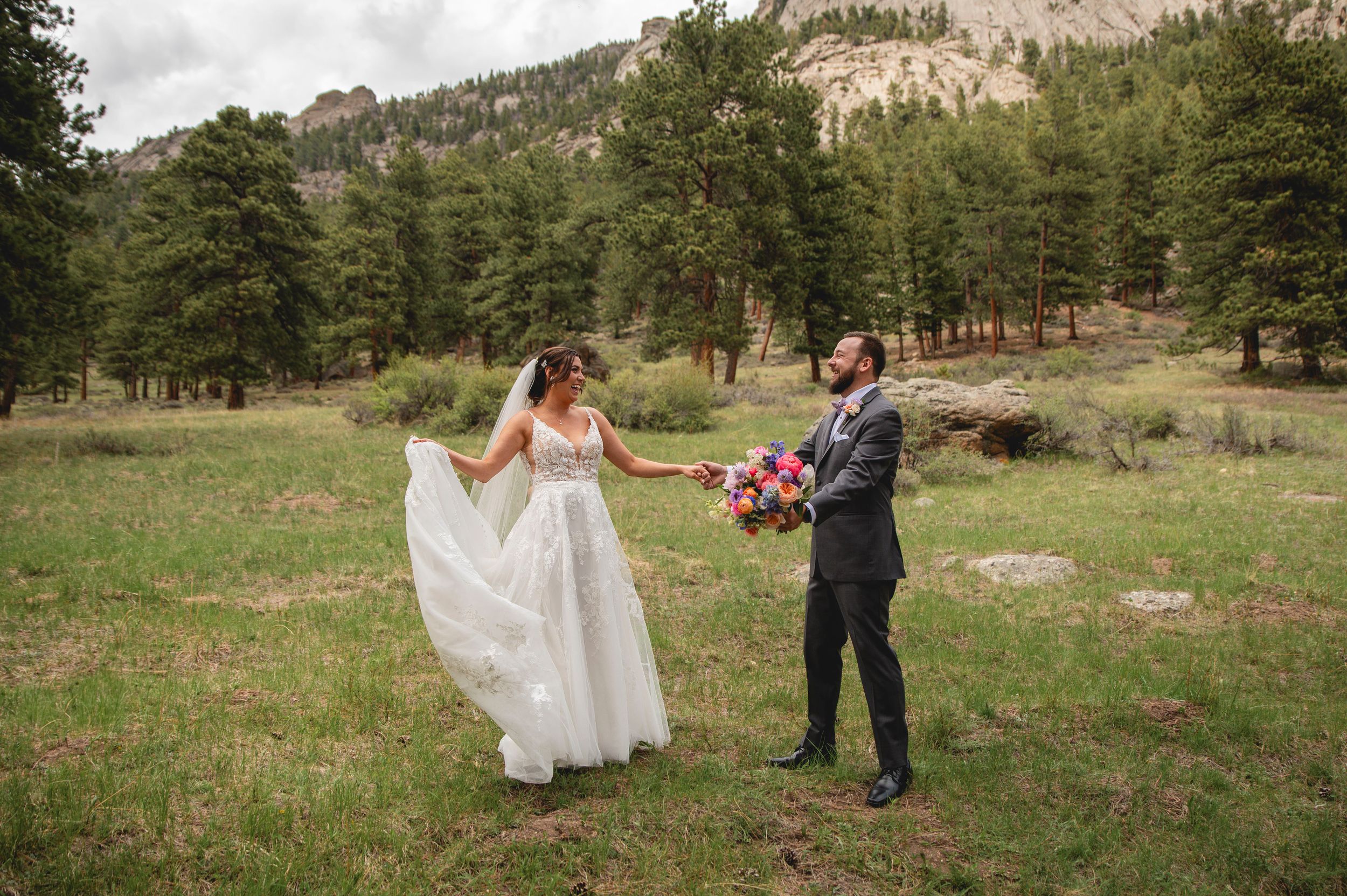 A couple in wedding attire stands in a scenic mountain meadow surrounded by pine trees and rocky cliffs.