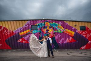 A couple in formal wedding attire poses in front of a vibrant street art mural with colorful geometric designs.