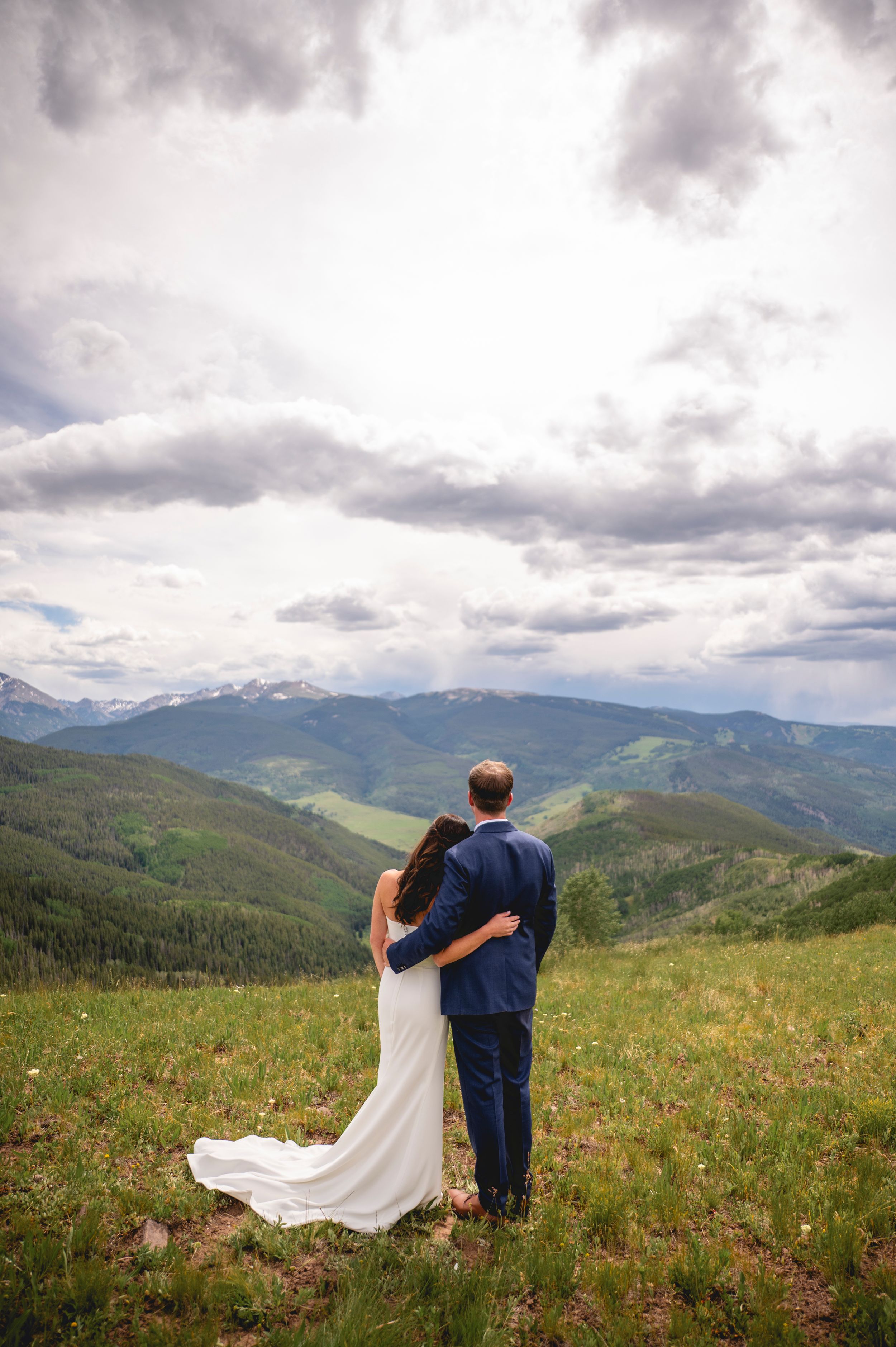 Bride and groom embrace while gazing at stunning mountain valley views under dramatic cloudy skies on their wedding day.