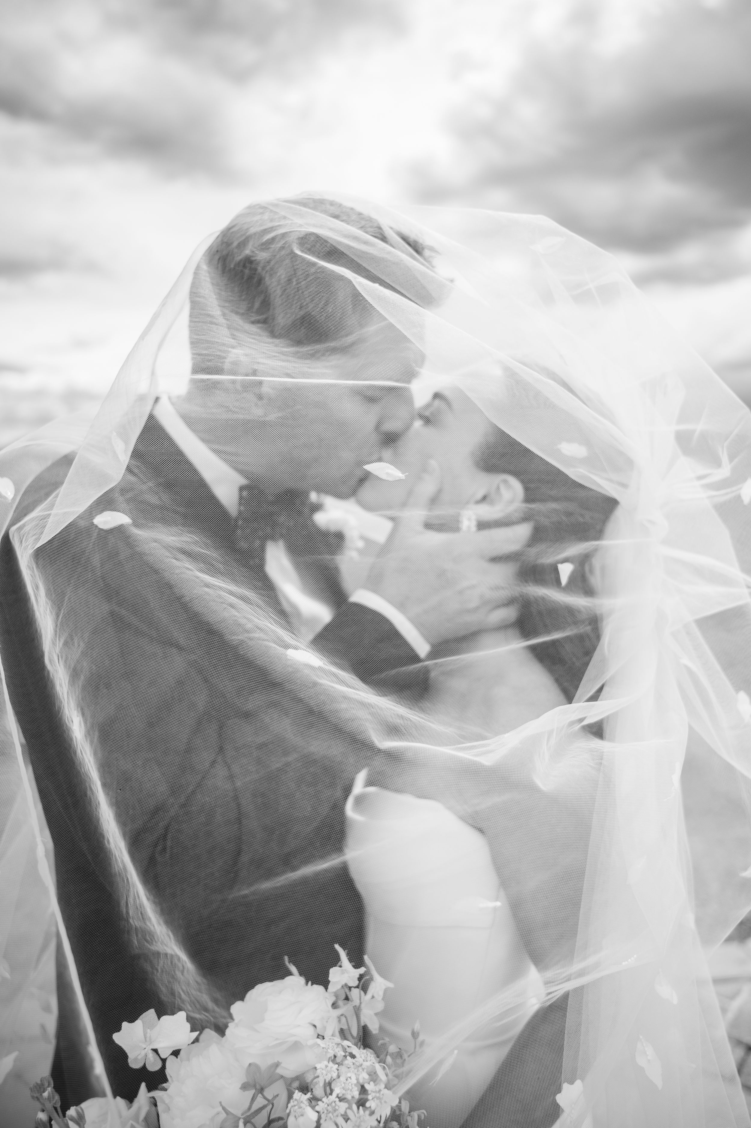Black and white photo of a bride and groom kissing under a flowing veil on their wedding day.