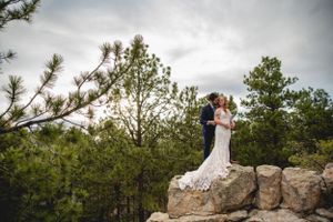 Wedding couple embraces on rocky mountain outcropping surrounded by pine trees.
