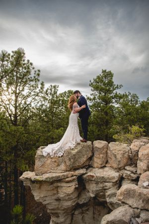 Wedding couple embracing on dramatic rocky cliff overlook with stormy skies.