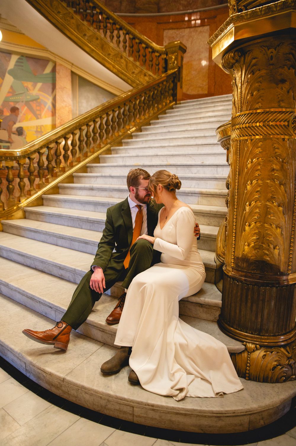 A couple in wedding attire sits romantically on marble stairs inside an elegant historic building with ornate gold columns.
