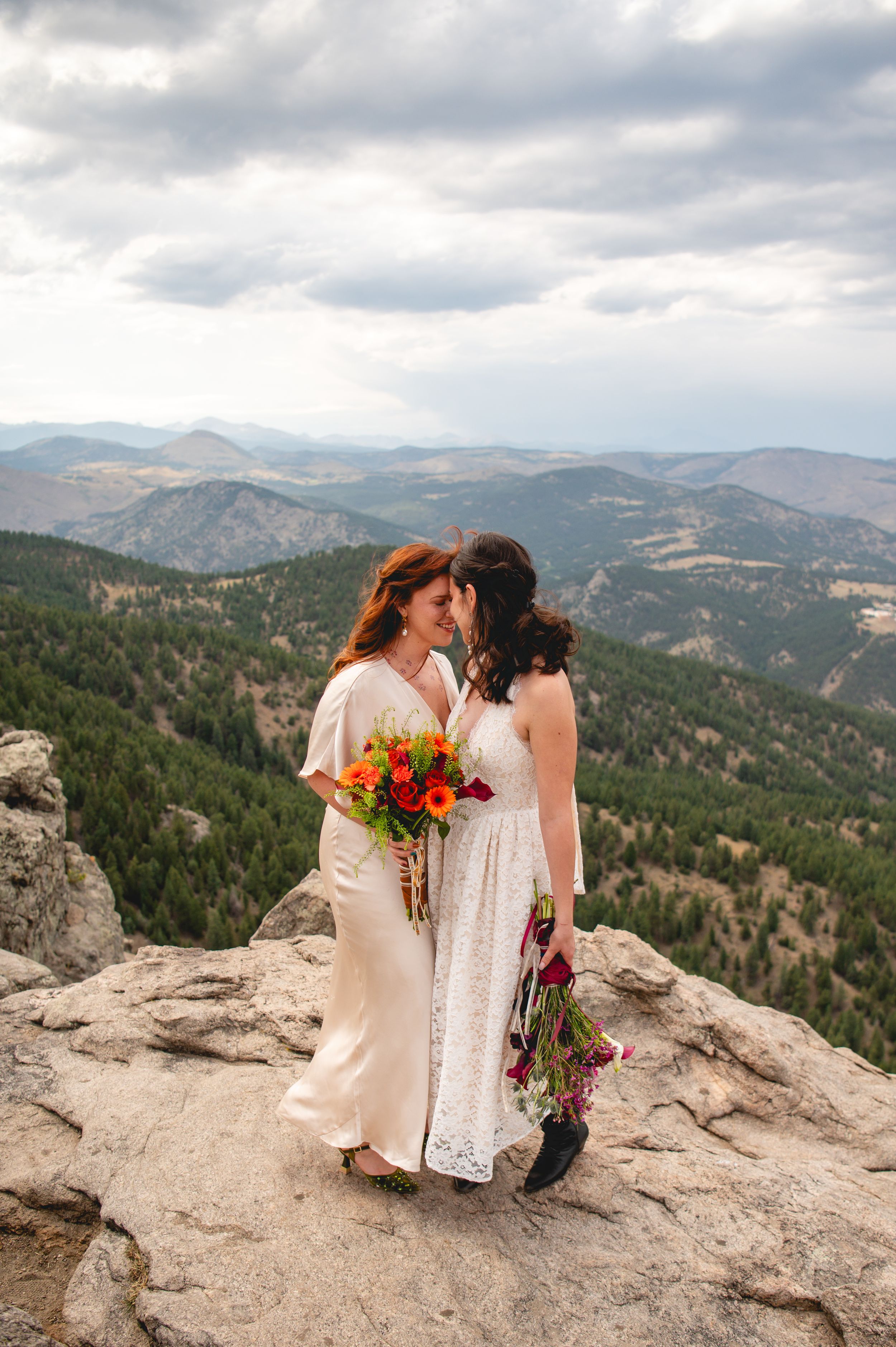 Two brides in white dresses share a romantic moment on a rocky mountain summit with sweeping Colorado landscape views.