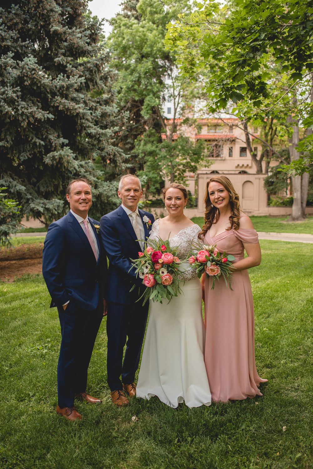 Small wedding group poses in formal attire on manicured lawn with historic building backdrop.