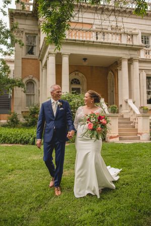 Wedding couple walking together on lawn in front of elegant historic mansion with white dress and blue suit.