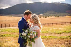 Couple poses with bouquet in field with mountain ranch backdrop.