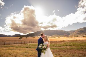 Wedding couple embraces in sunny mountain valley with dramatic clouds and ranch fencing.