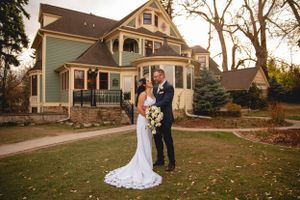 A couple in wedding attire poses on the front lawn of a beautiful Victorian style sage green house at sunset.