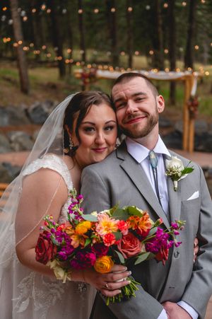 Bride and groom pose together outdoors, she holds vibrant bouquet of pink and orange flowers, forest and lights in background.