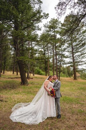 Bride and groom embrace in a pine forest clearing, her lace gown trailing elegantly, holding a vibrant bouquet.