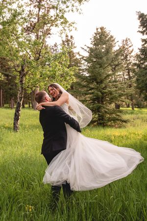 A romantic wedding photo of a couple dancing in a lush green field surrounded by evergreen trees and birch trees.