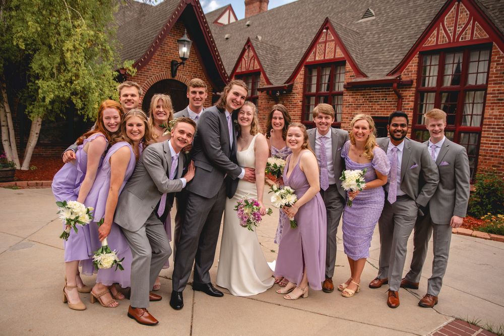 Wedding party in purple and gray attire pose together outdoors in front of rustic Tudor-style building.