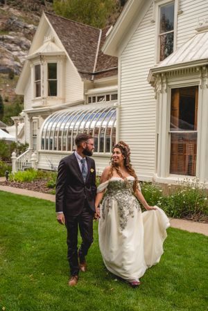 Wedding couple strolling on lawn beside white Victorian house with glass conservatory.