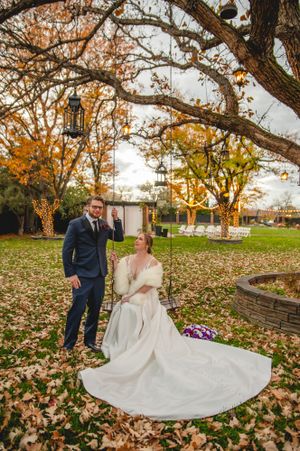A couple takes wedding photos in an autumn garden setting with fallen leaves and a tree swing on a cloudy day.