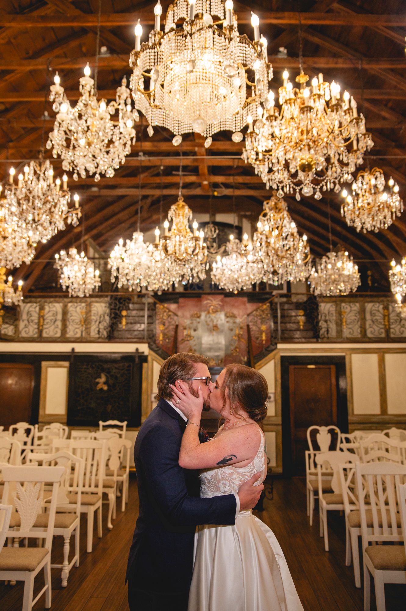 Ornate crystal chandeliers illuminate a grand wedding ceremony venue with white chairs.