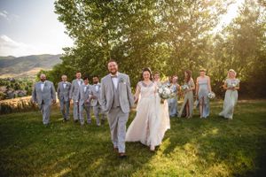 Wedding party walks across green hillside in light gray suits and white gowns.