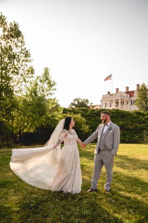Couple poses in formal wear on a green lawn with historic mansion and American flag in background.