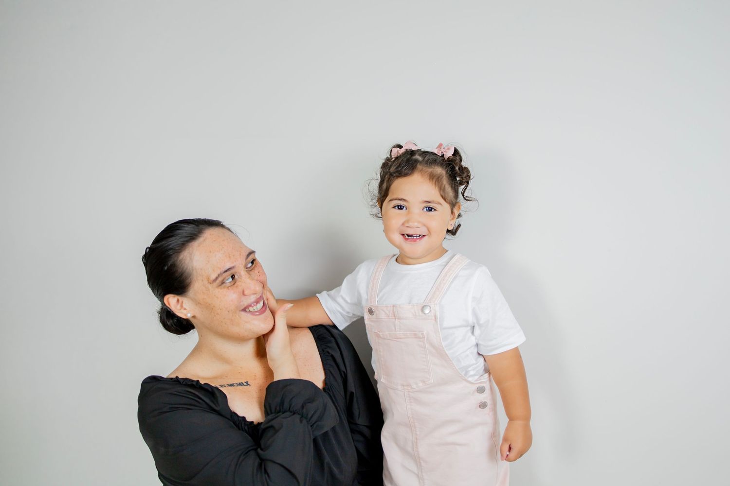 Smiling mother in black top holds happy toddler girl in pink overalls against white background.