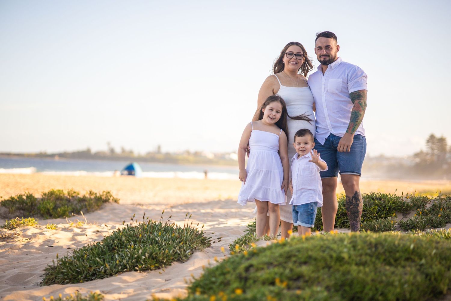 Family of four in white outfits posing together on a sunny beach with ocean in background.