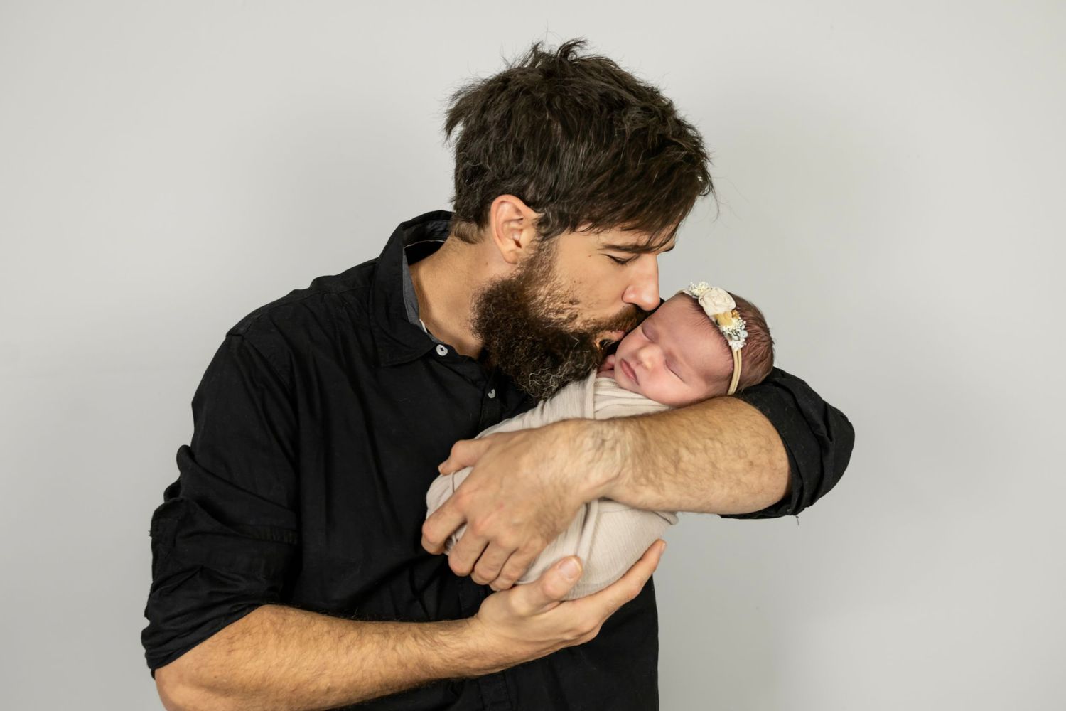 Bearded father in black shirt gently kisses swaddled newborn baby in studio portrait.