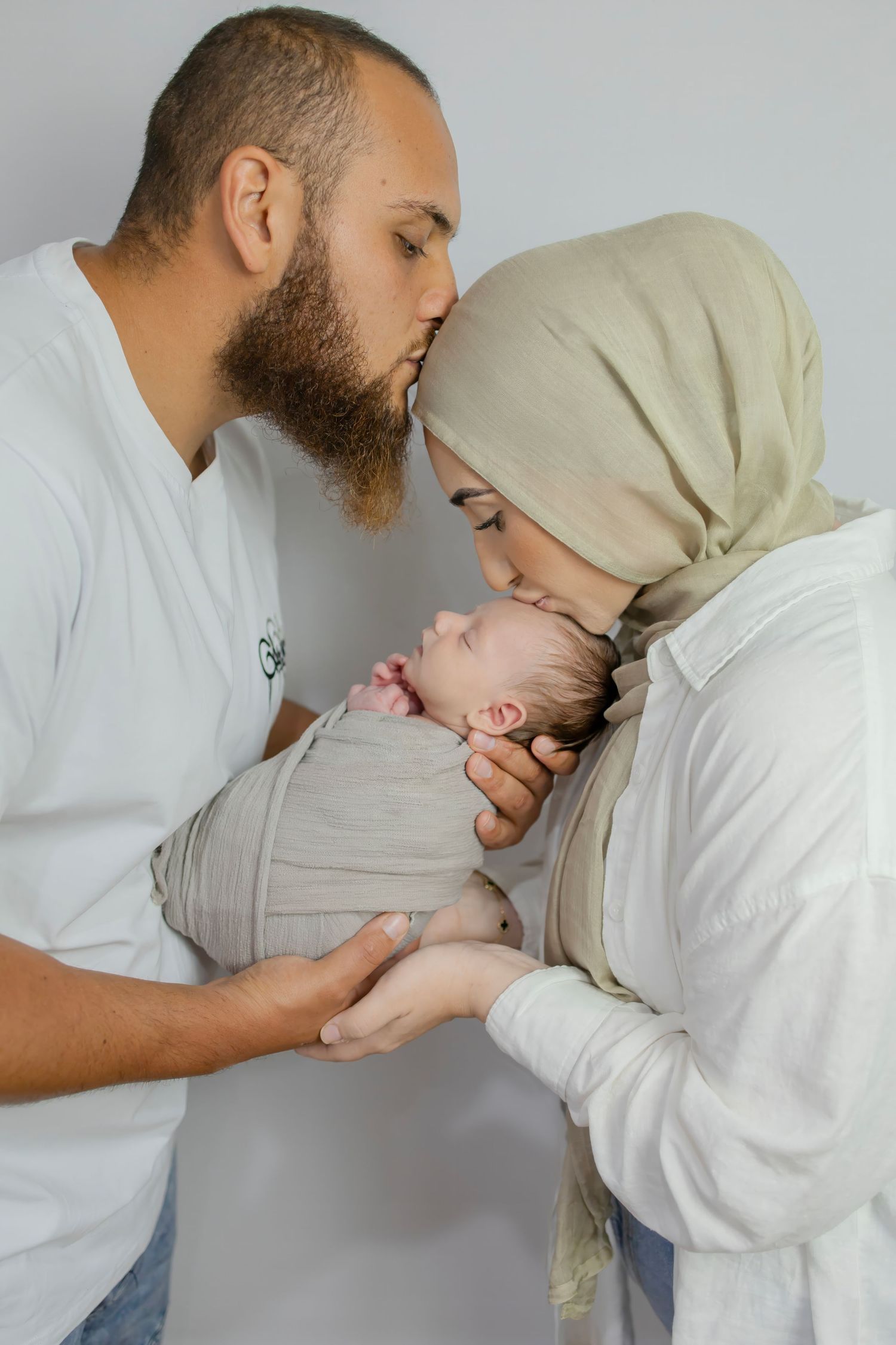 Muslim parents in white outfits kiss their sleeping swaddled newborn baby together.