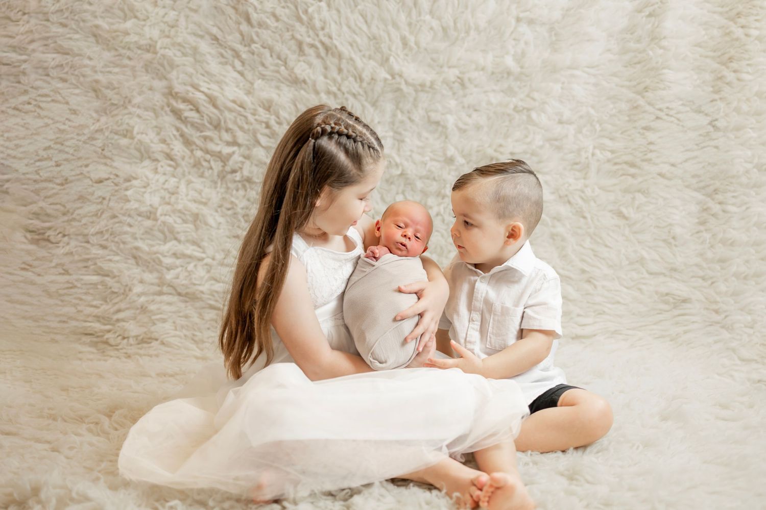 Two siblings in white outfits sit on fluffy rug sharing newborn baby between them.
