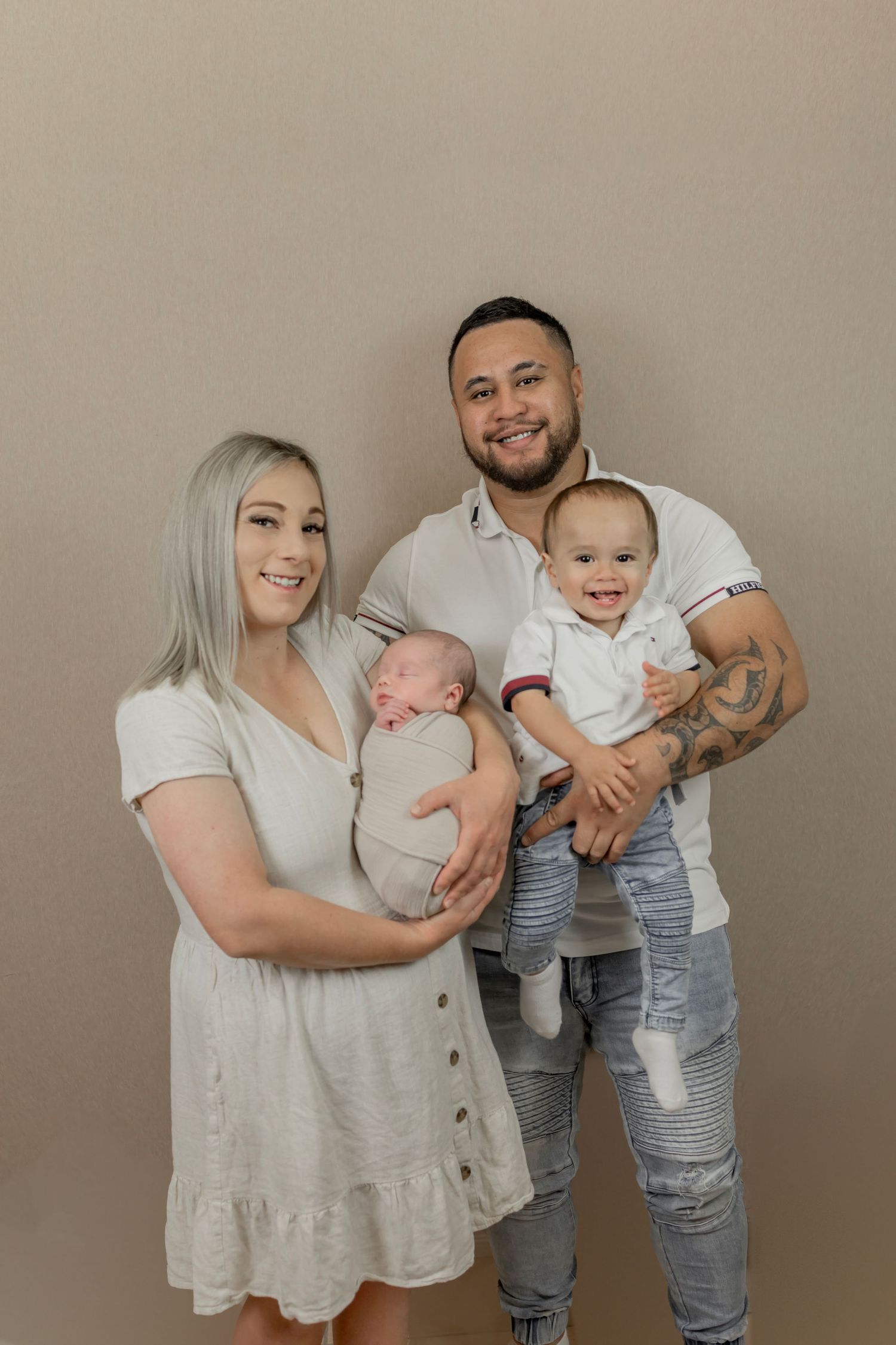 Young family portrait with mother holding newborn baby and father carrying toddler son against neutral background.