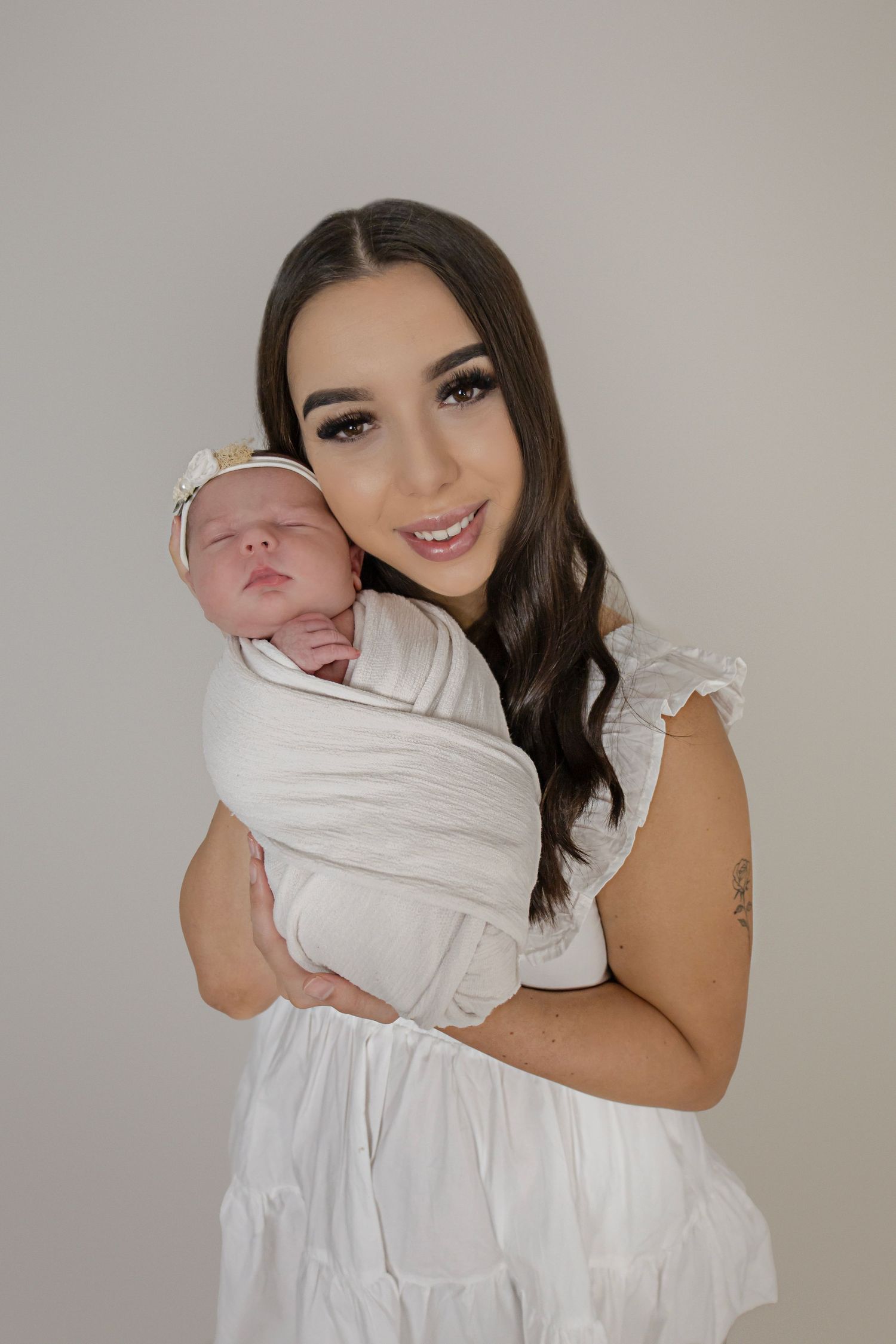 Young mother in white dress holds swaddled newborn baby with pink headband in professional studio portrait.