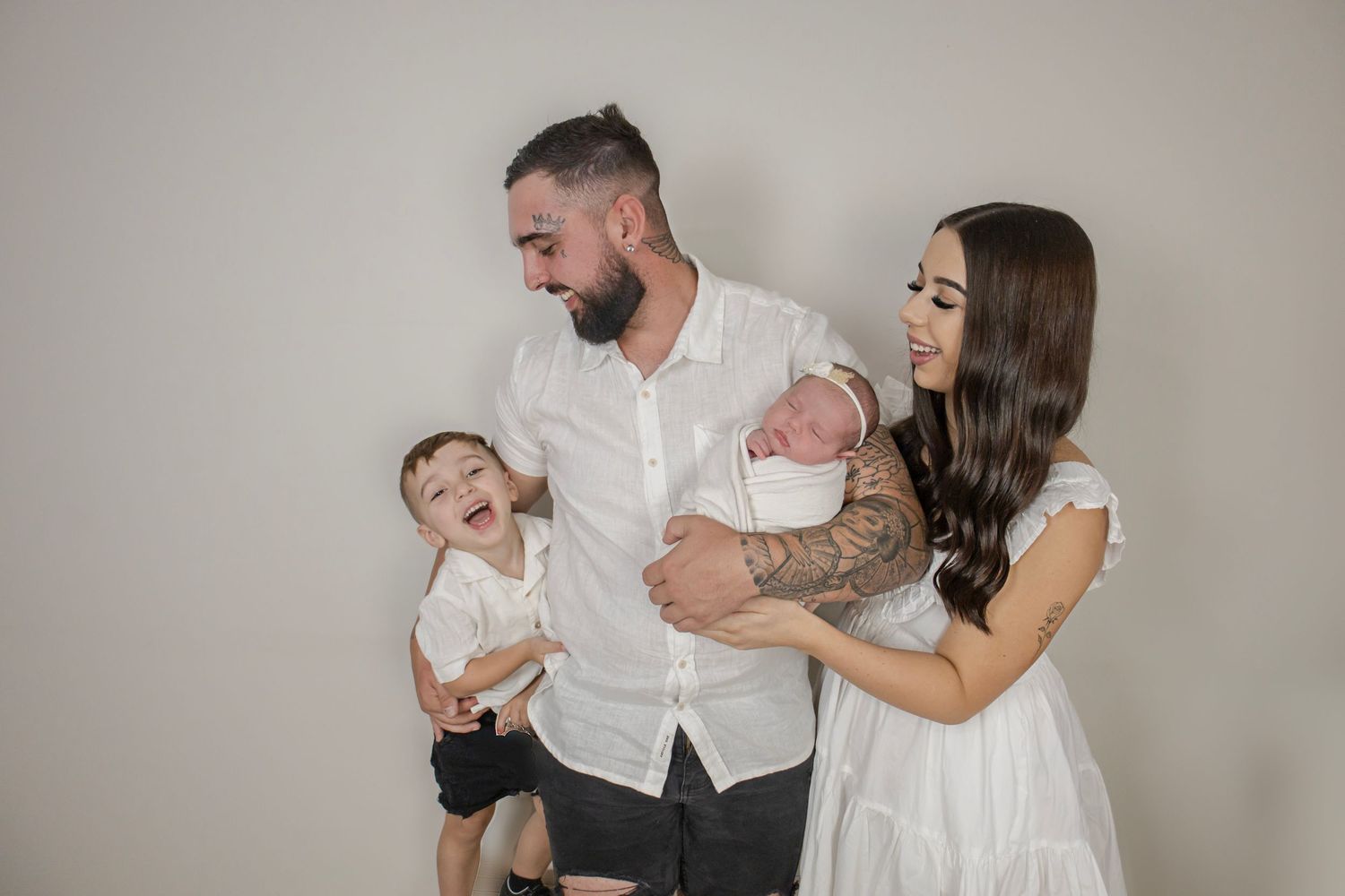 Tattooed couple with laughing toddler and newborn baby in white studio setting.