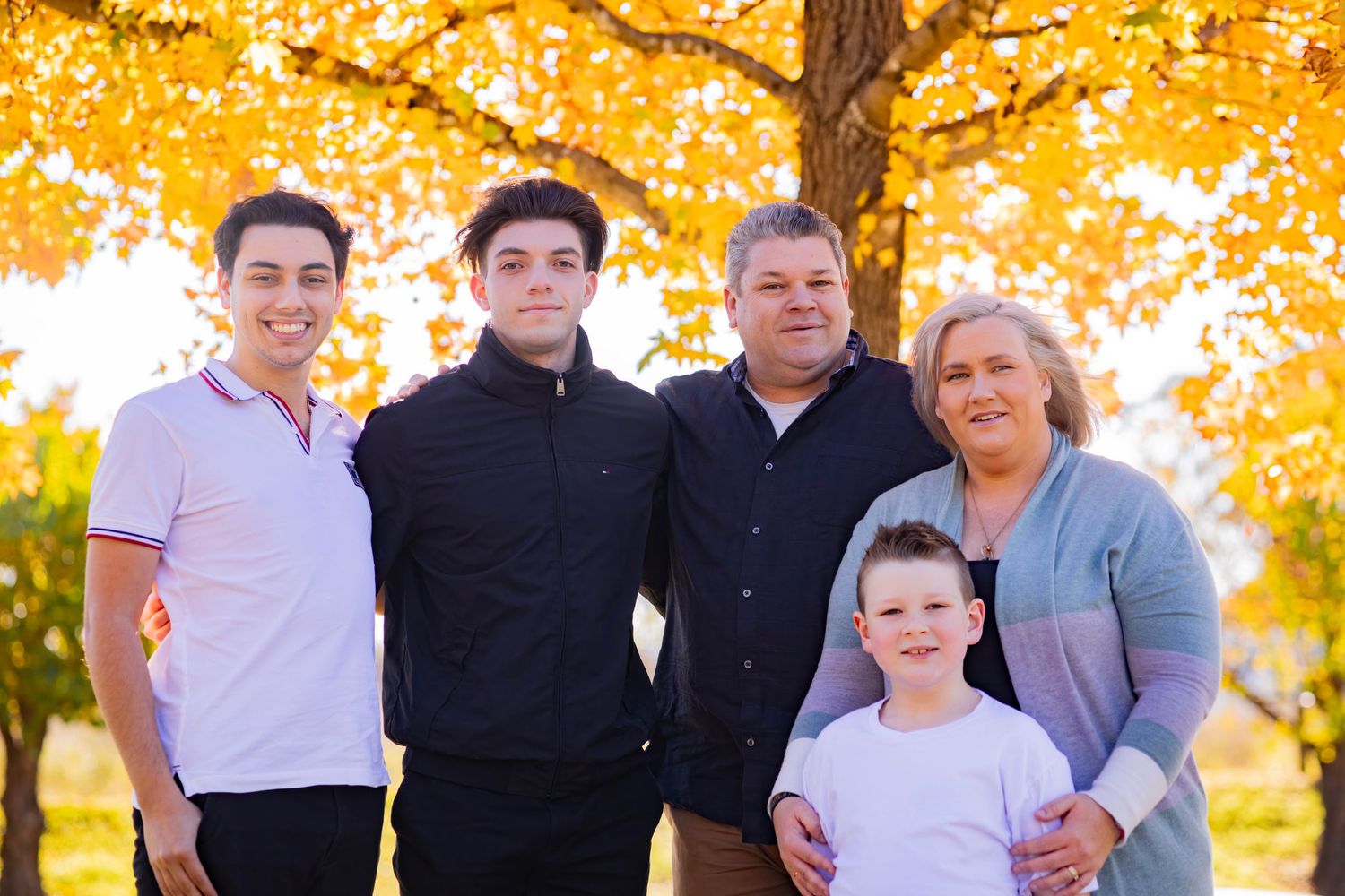 Family of five with three sons and parents smiling together under golden autumn foliage outdoors.