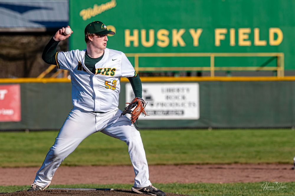 Baseball pitcher in green and gold uniform mid-throw at Husky Field during a sunny game.
