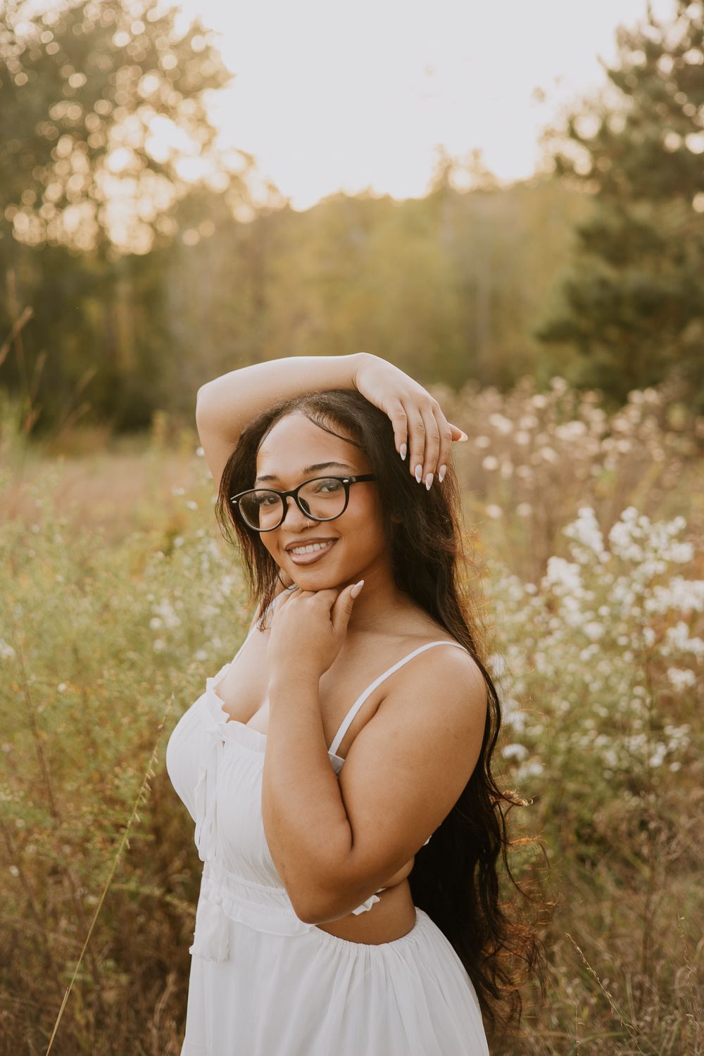 Woman in glasses and white dress poses playfully in sunlit meadow.