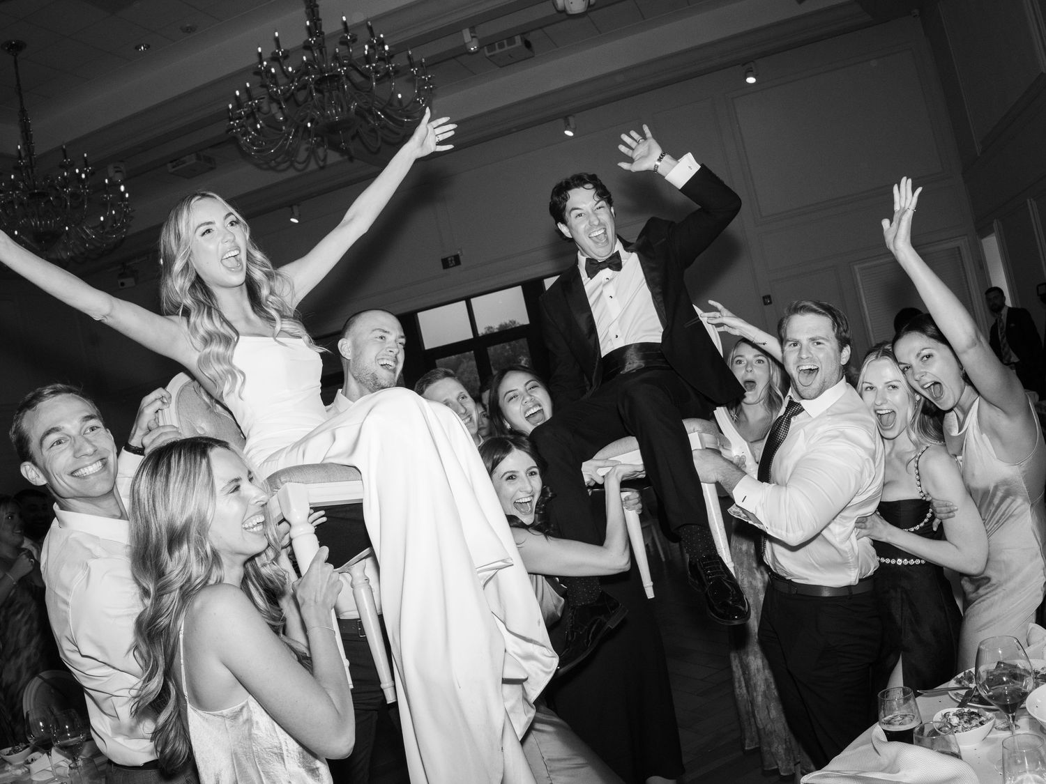 Wedding reception guests dancing and celebrating with arms raised on dark dance floor.