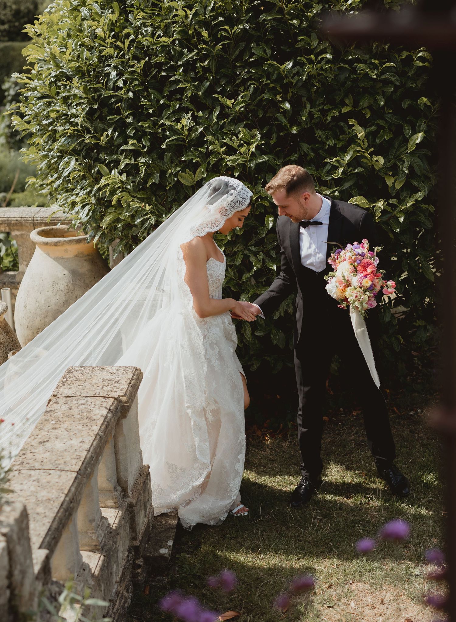 A romantic wedding scene with a bride in a flowing veil and groom holding a pink bouquet in a garden setting.
