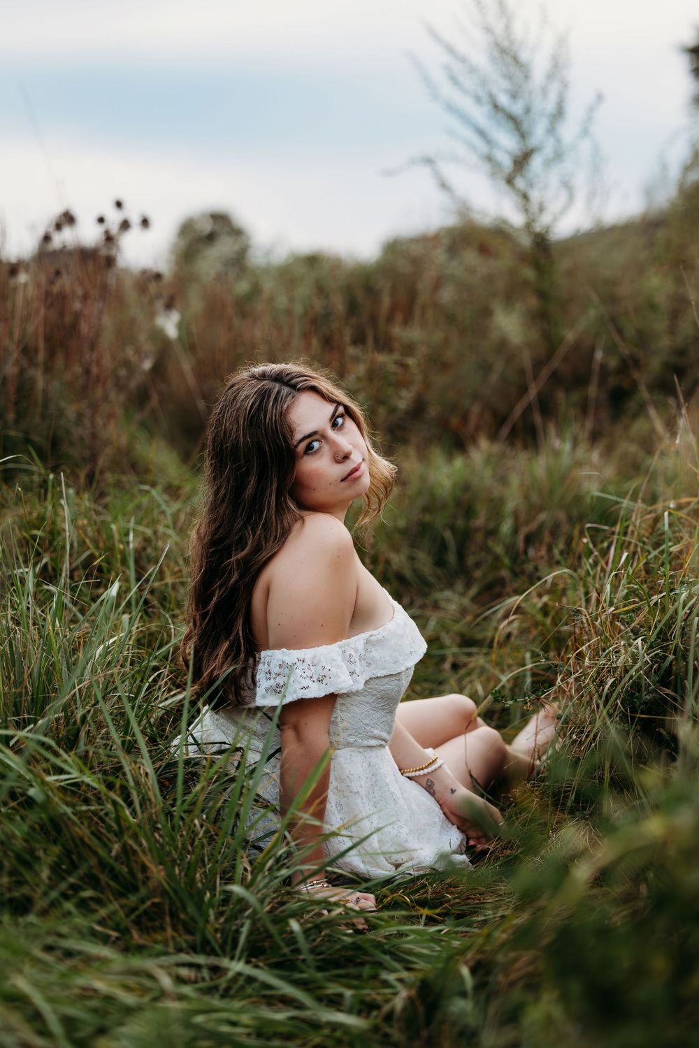 High school senior portrait in white off-shoulder dress poses gracefully among tall grass in natural setting.