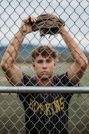 Teen baseball player in Hopkins jersey holds glove above chain-link fence outdoors.