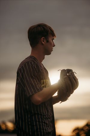 Young baseball player in pinstripe jersey holds mitt at sunset, silhouetted against dramatic sky.