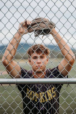 Teen baseball player in Hopkins jersey holds glove above chain-link fence on outdoor field.