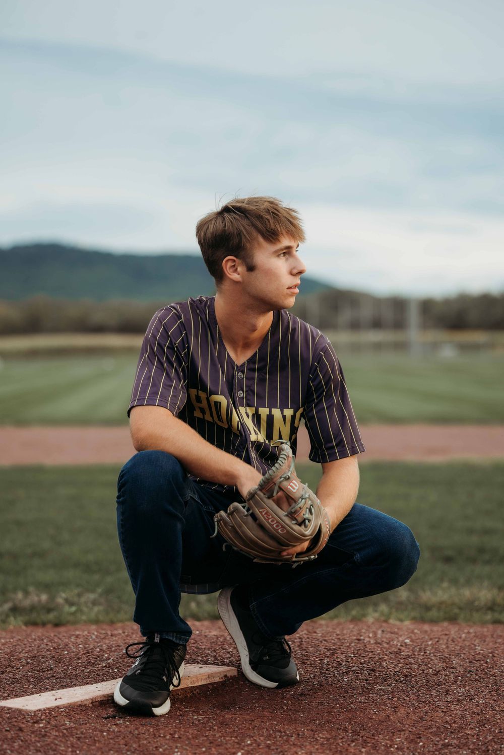 Young baseball player crouching on base wearing jersey and glove on an outdoor field.