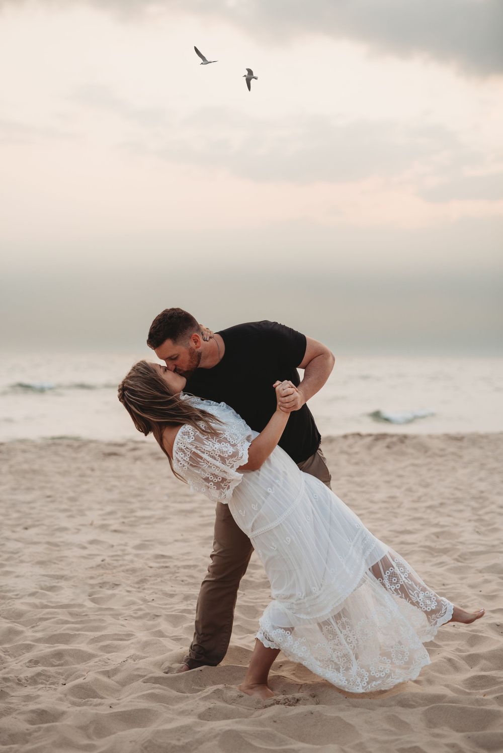 Couple sharing romantic dip kiss on sandy beach with seabirds flying overhead.