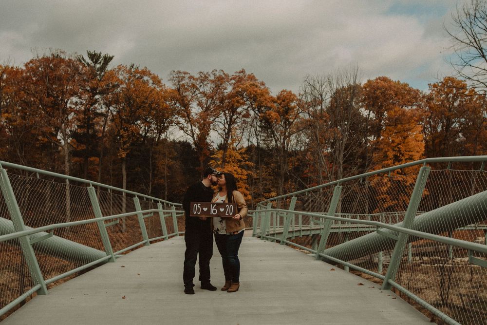 Couple standing on modern bridge walkway surrounded by autumn foliage.