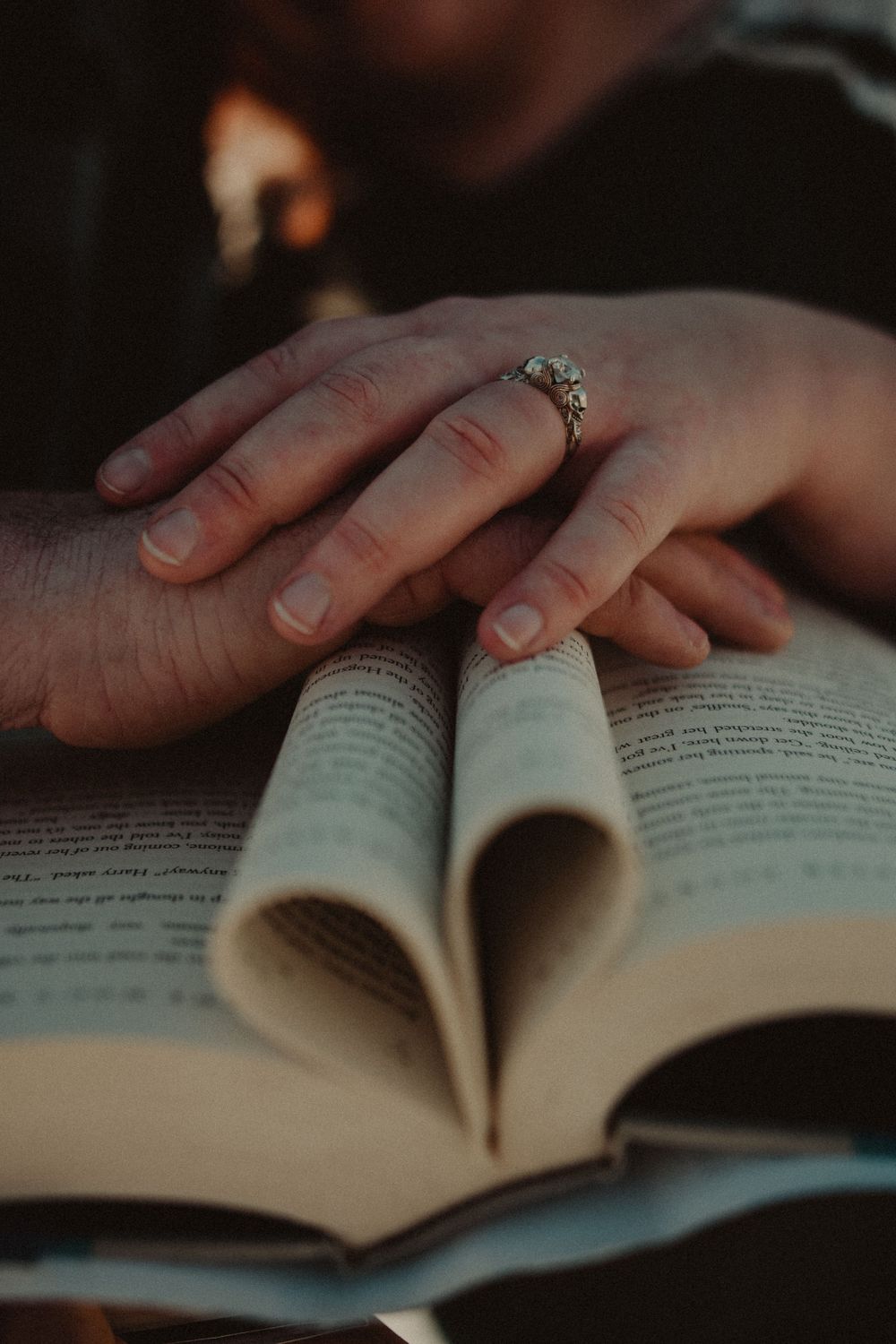 Hands resting on an open book with pages forming a heart shape, featuring an engagement ring.