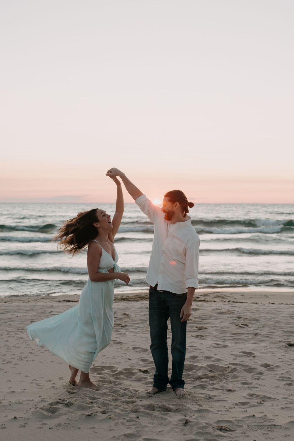 A couple dances playfully on the beach during sunset with waves crashing behind them.