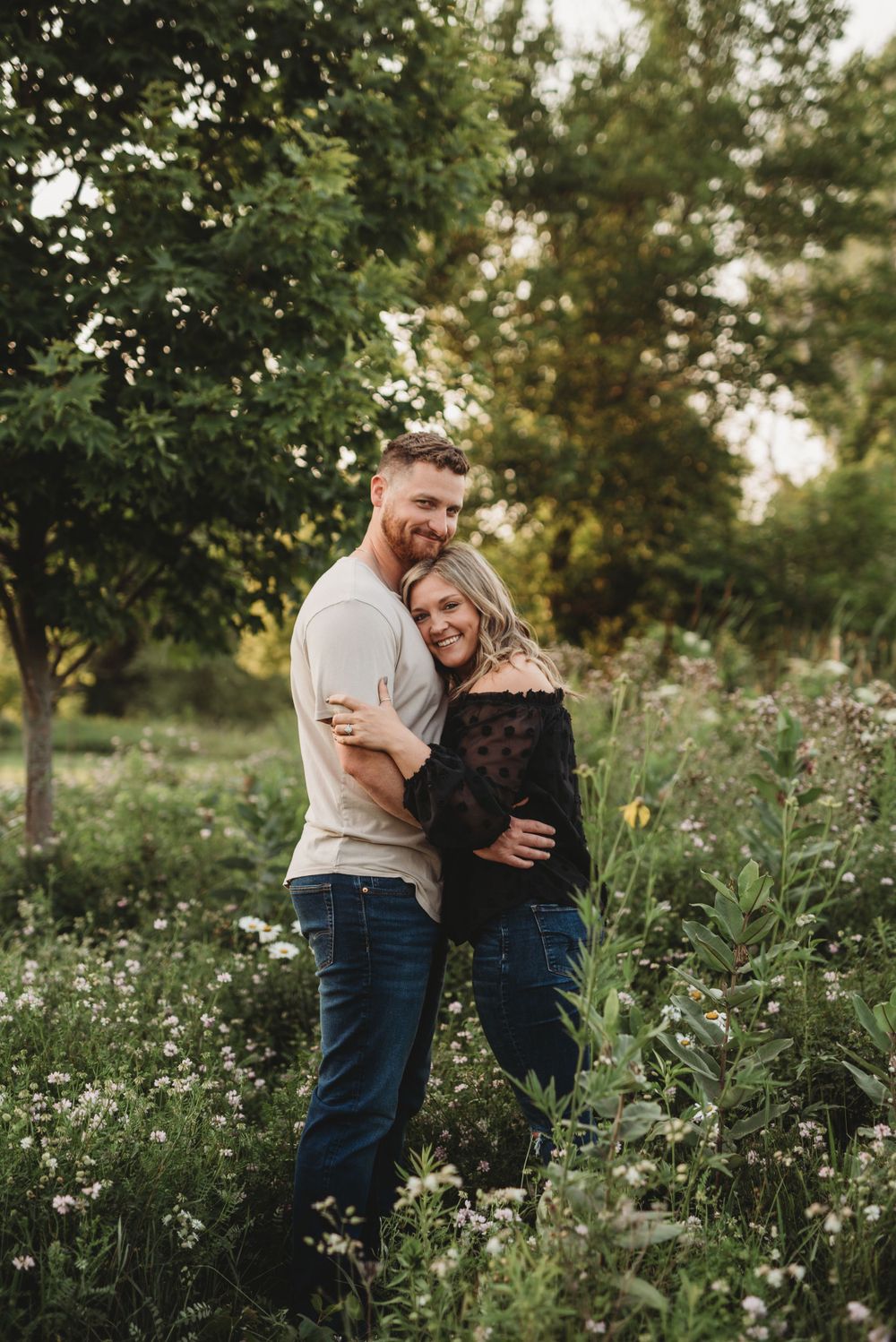 A couple embraces in a flowering meadow with golden sunlight filtering through the trees.