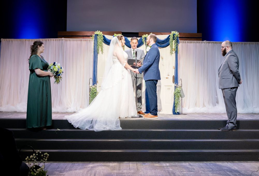 Wedding ceremony on stage with bride, groom, officiant, and bridesmaid under floral arch.
