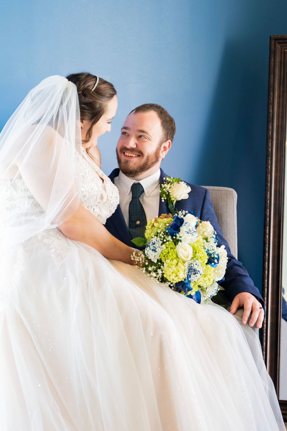 Smiling groom in navy suit looks at bride holding blue and yellow bouquet while seated together.