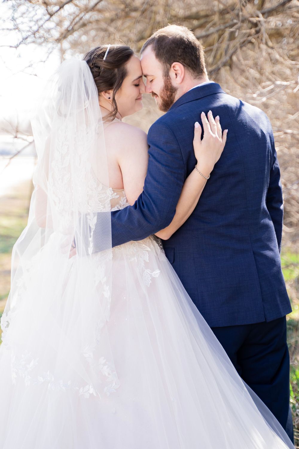 Bride and groom touch foreheads tenderly in an outdoor wedding portrait with lace dress and veil.