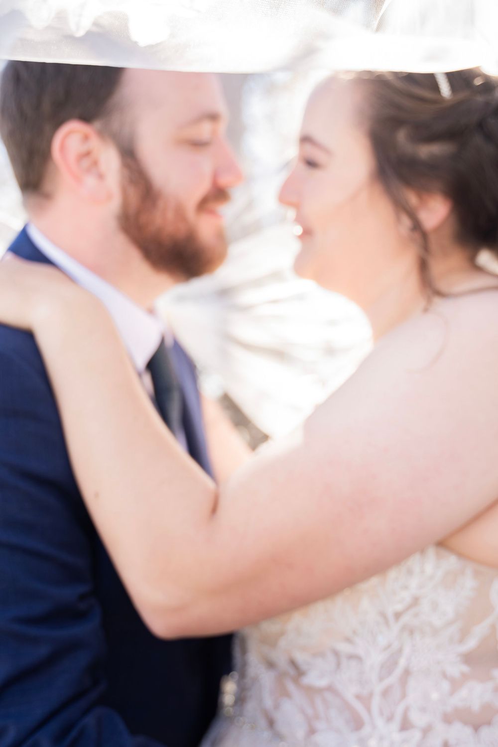 Soft focus romantic portrait of bride and groom embracing closely beneath a flowing white veil.